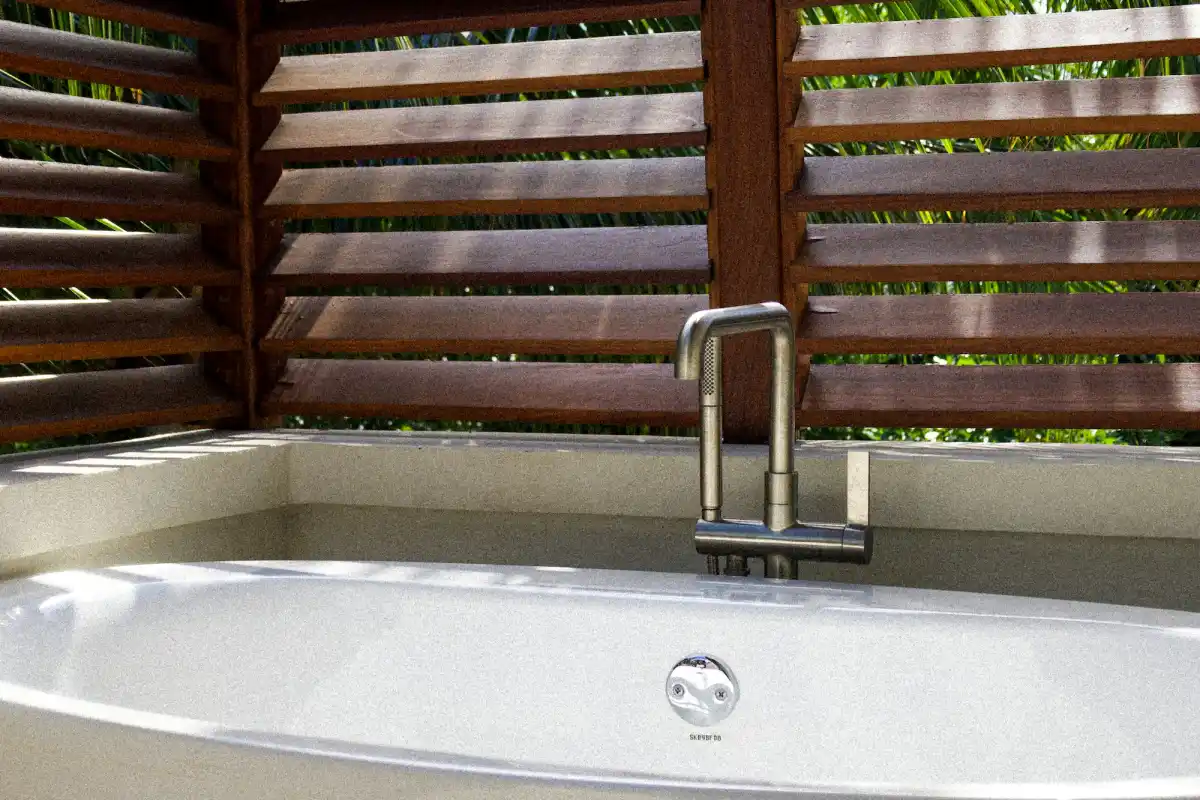 Bồn tắm trắng đặt sát vách lá sách gỗ, nắng lọt qua khe